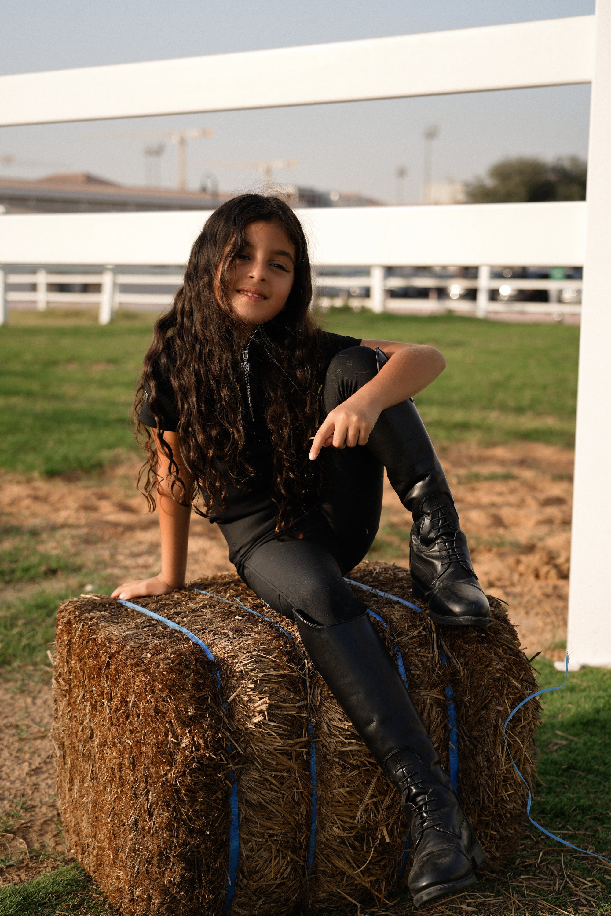 Person sitting on a hay bale in an outdoor setting with a white fence and grass in the background.